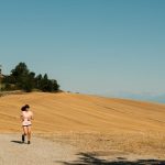 A woman runs on a gravel path through a sunlit rural landscape with dry fields and blue sky.